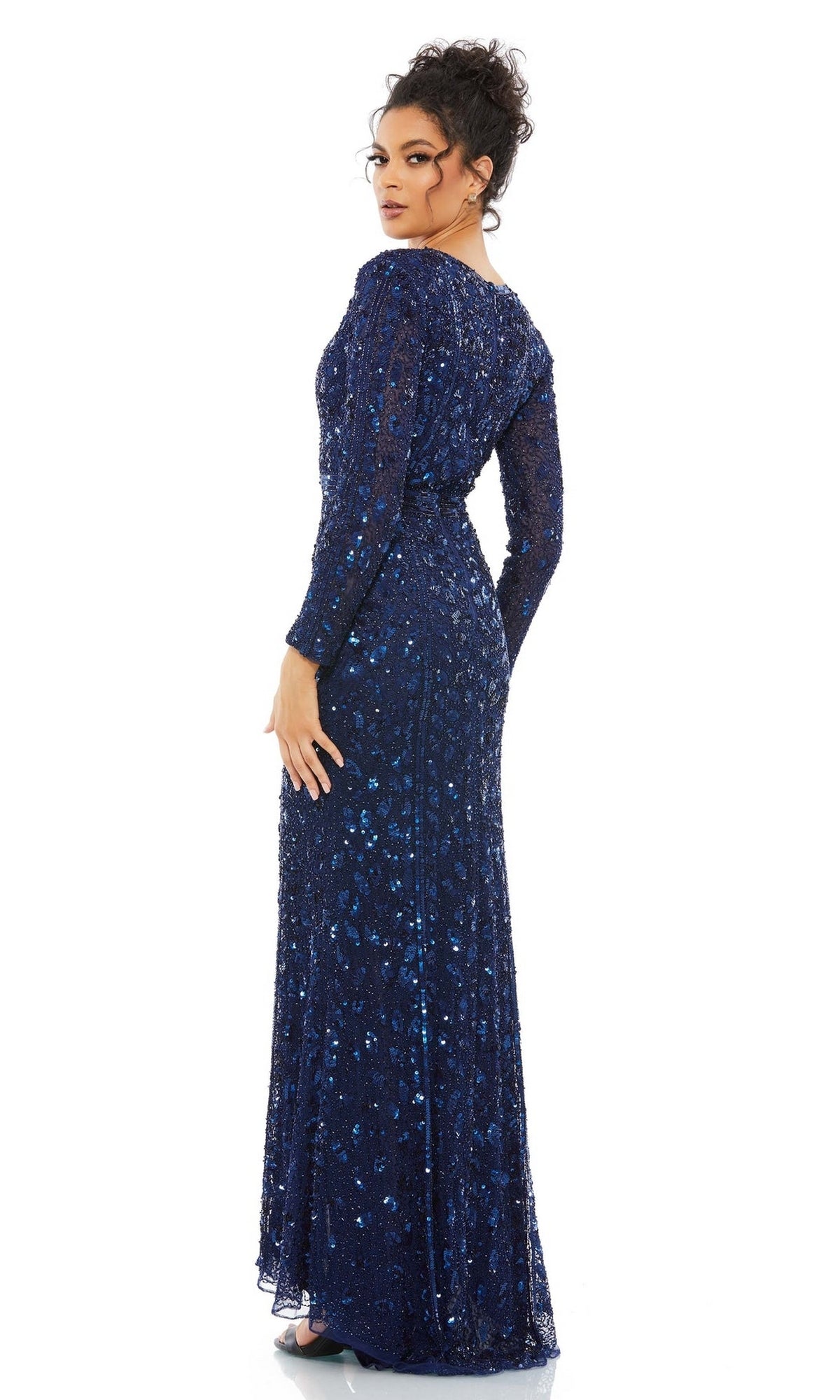 A woman stands facing slightly away, wearing the Long Formal Dress 5002 by Mac Duggal—a floor-length, dark blue sequin gown with long sleeves, a fitted waist, and textured fabric—set against a plain white background.